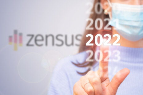 Frau mit Mund-Nasen-Schutz zeigt auf Jahreszahl 2022, Zensus-Logo im Hintergrund sichtbar.